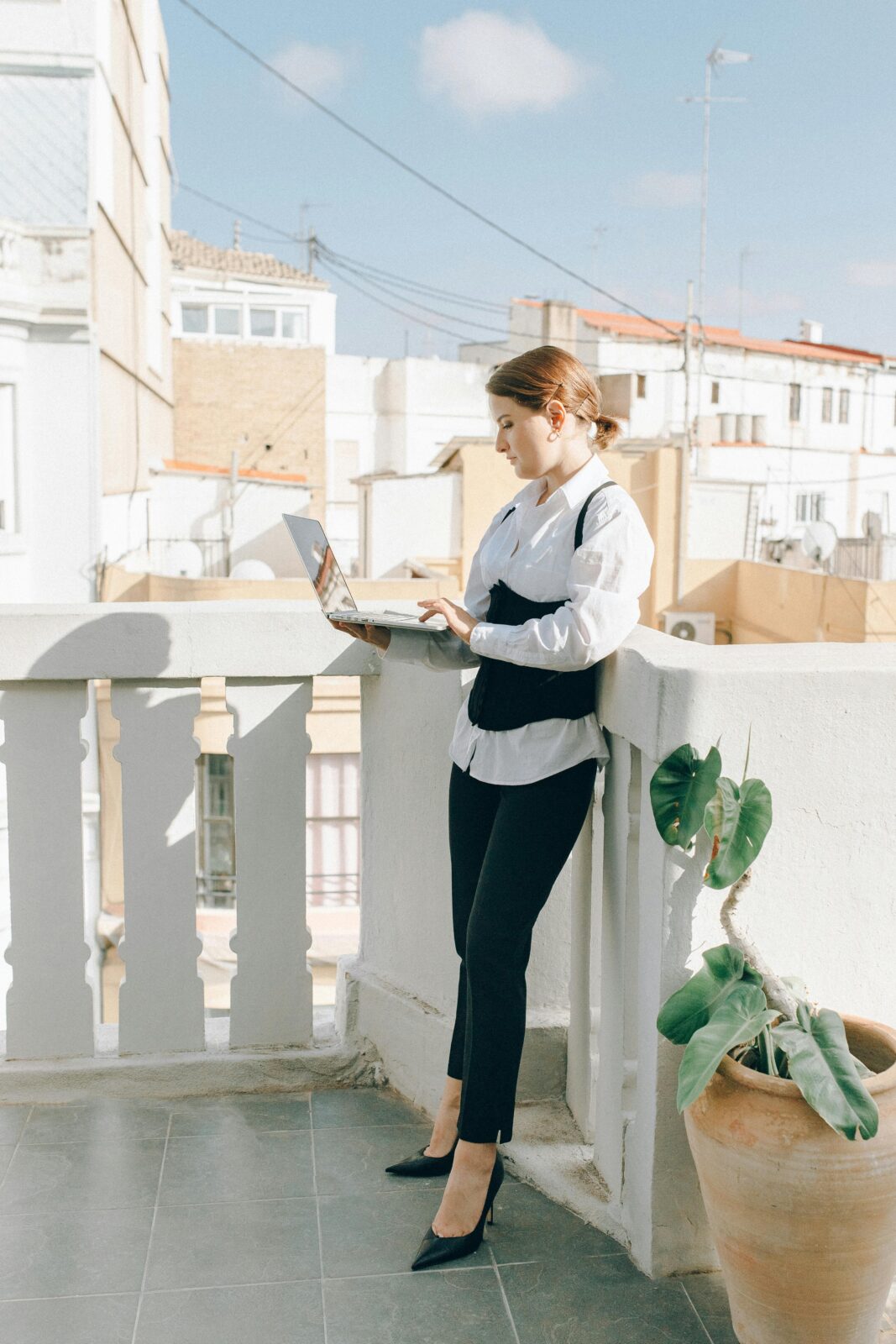 A businesswoman standing on an outdoor balcony using a laptop in corporate attire.